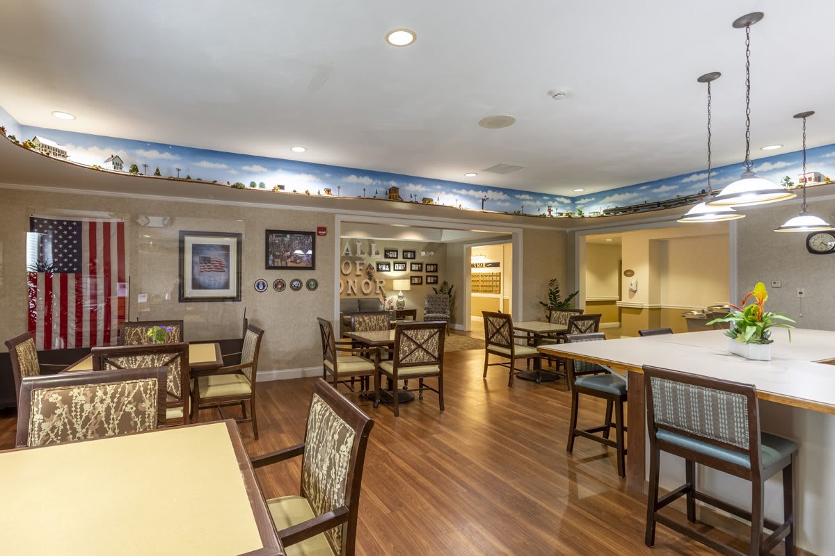 A cozy dining area with tables and chairs set on a wooden floor, featuring artwork on the walls and warm lighting.