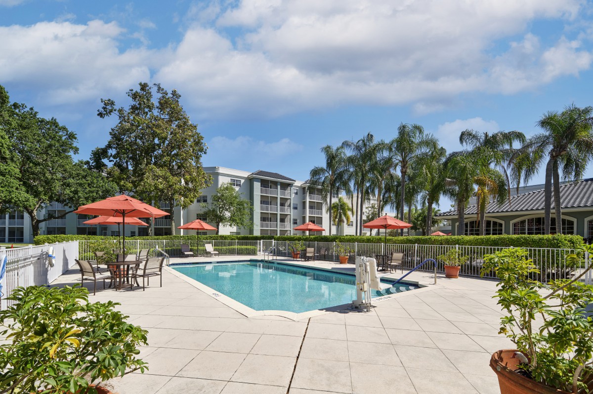 An inviting outdoor swimming pool area surrounded by palm trees and residential buildings, featuring lounge chairs and umbrellas for relaxation.