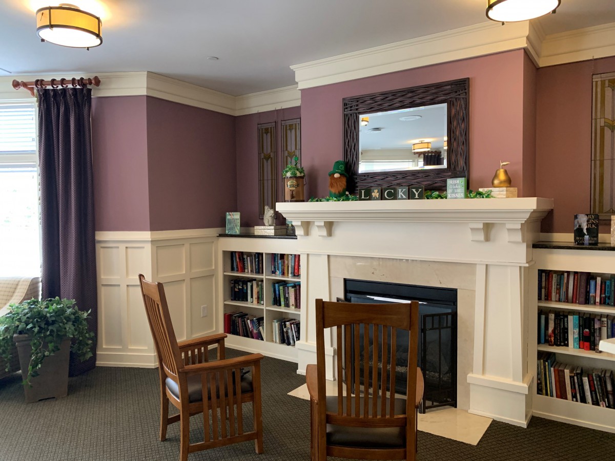 A cozy interior room featuring a fireplace, decorated with a mirror and bookshelf, and equipped with wooden chairs.