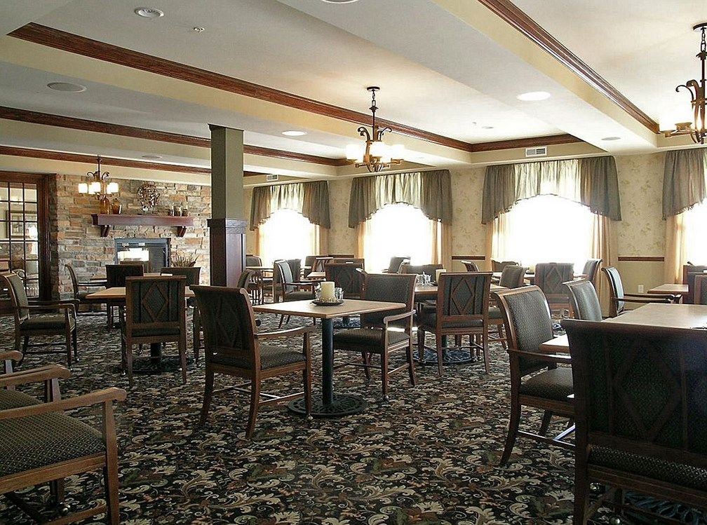 An empty dining area with tables and chairs arranged neatly, featuring large windows and a patterned carpet.
