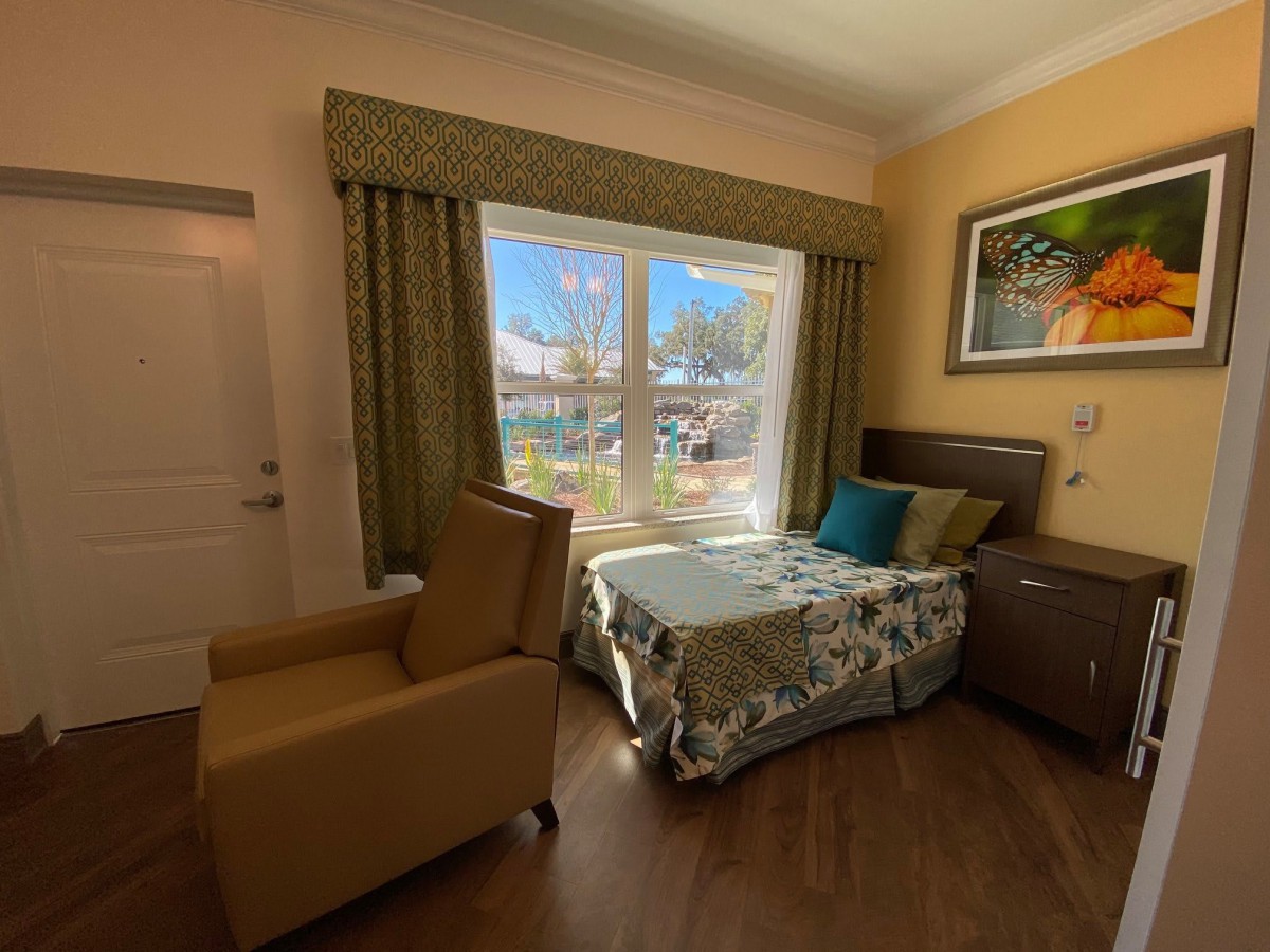 A well-lit hospital room with a bed, chair, and a large window with curtains.