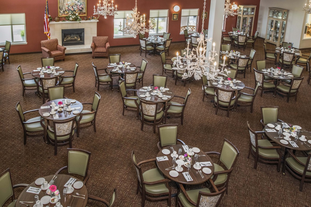 An elegant dining room with multiple round tables neatly set for guests, featuring chandeliers and a warm decor.
