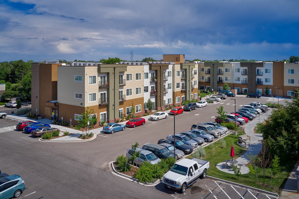 A modern apartment complex with a large parking lot and several parked cars under a cloudy sky.