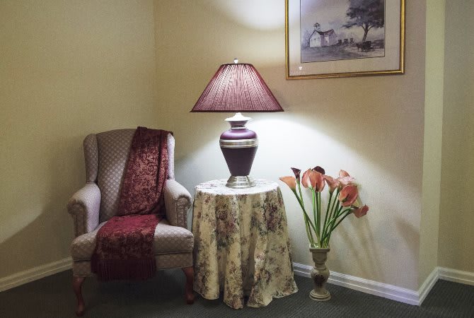 A cozy corner featuring an upholstered chair, a table with a decorative lamp, and a flower arrangement.