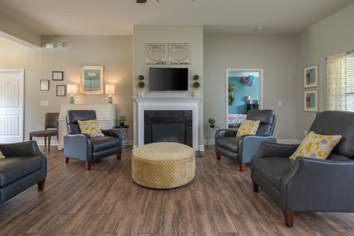 A cozy living room featuring gray armchairs, a central yellow ottoman, and a fireplace with a mounted television.