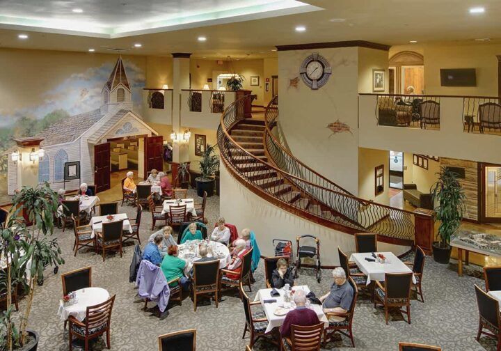 A spacious dining area with seniors gathered around tables, featuring a grand staircase and cozy ambiance.