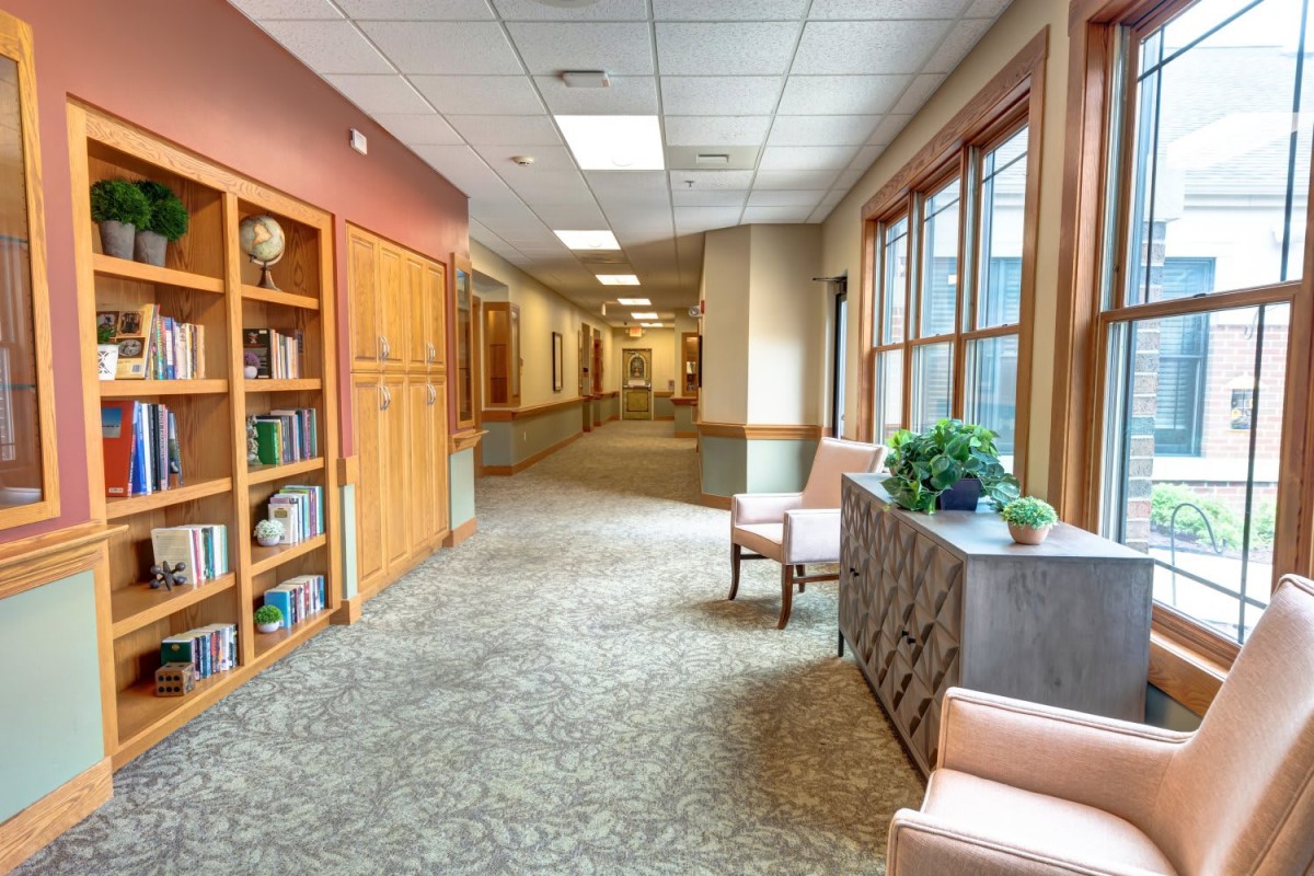 A quiet hallway with a seating area and bookshelf, featuring large windows and indoor plants.