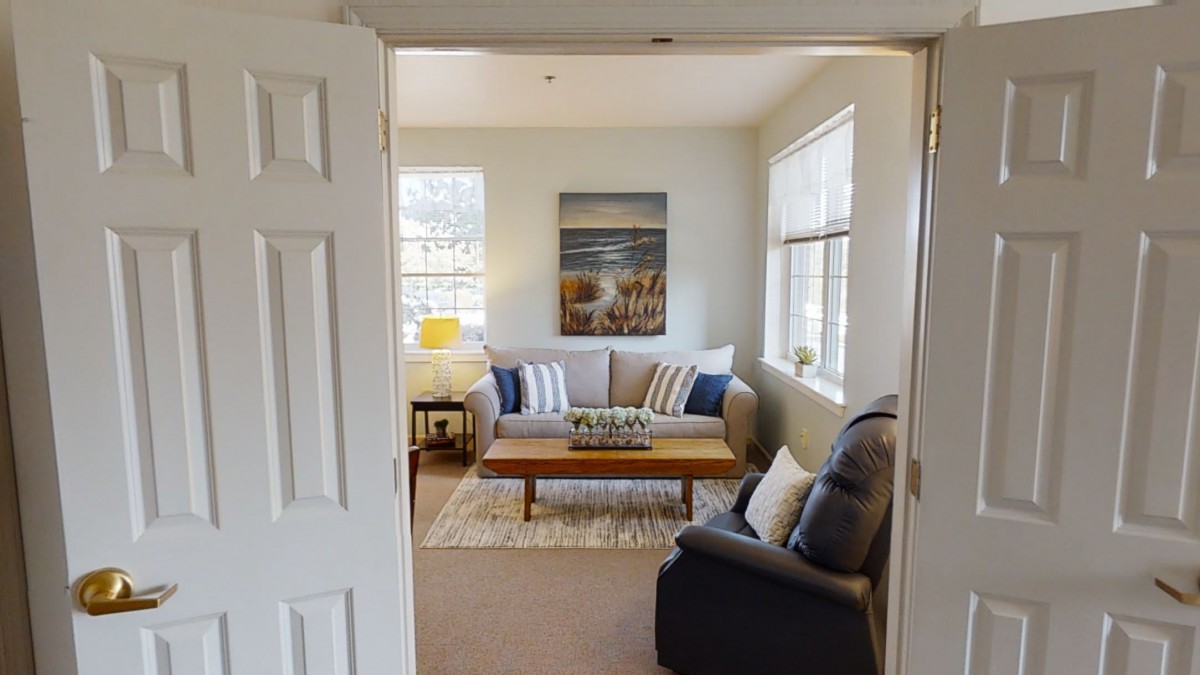 A cozy living room featuring a comfortable sofa and armchair, centered around a wooden coffee table. Large windows allow natural light to fill the space, while decorative pillows and wall art add a touch of style.