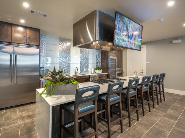 A modern kitchen with a sleek countertop, bar stools, stainless steel appliances, and a wall-mounted television.