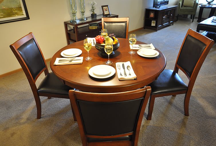 A round dining table set for four with neatly arranged plates, utensils, and glasses, featuring a fruit bowl centerpiece.