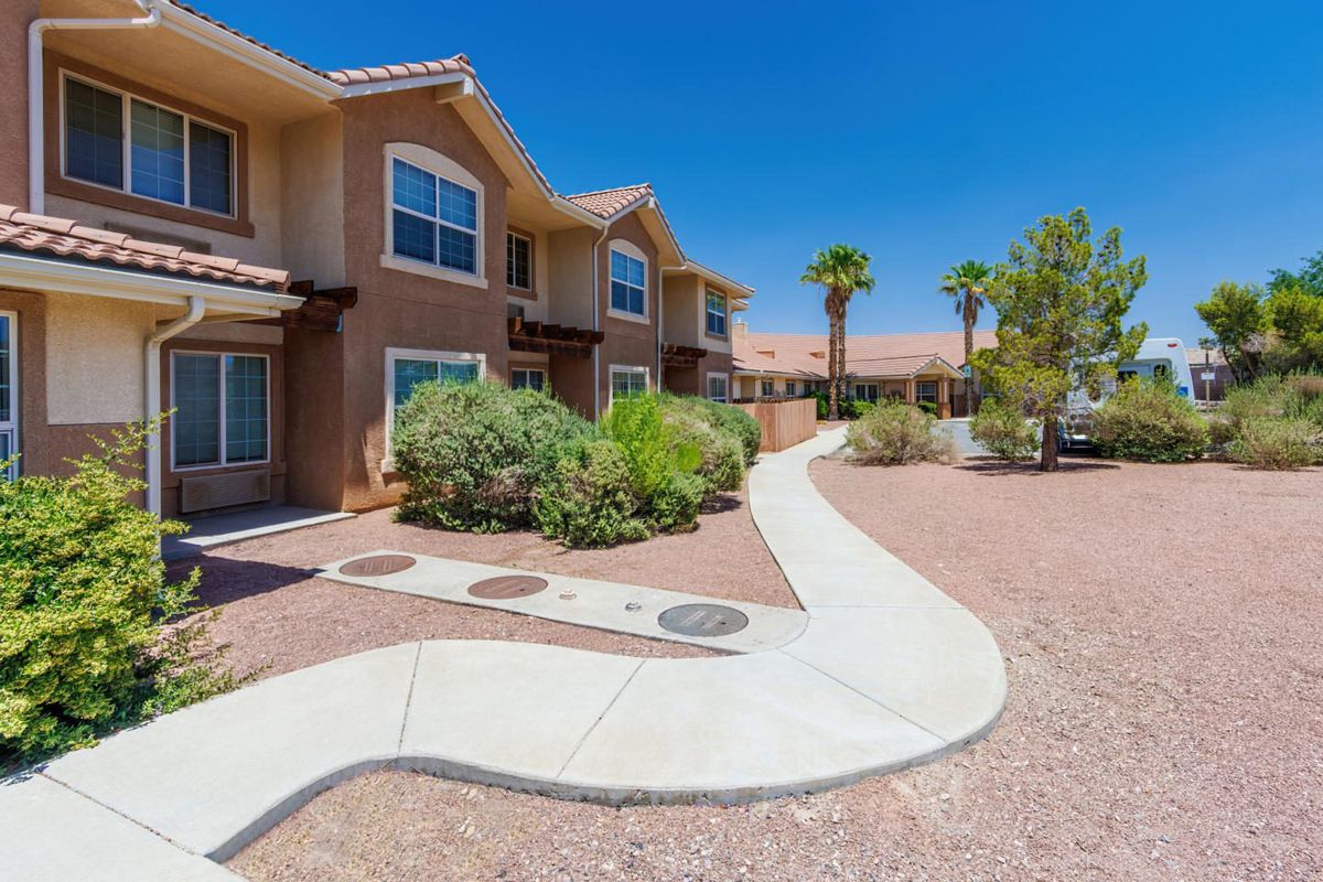 A row of residential apartments with a landscaped walkway surrounded by desert plants and palm trees on a sunny day.