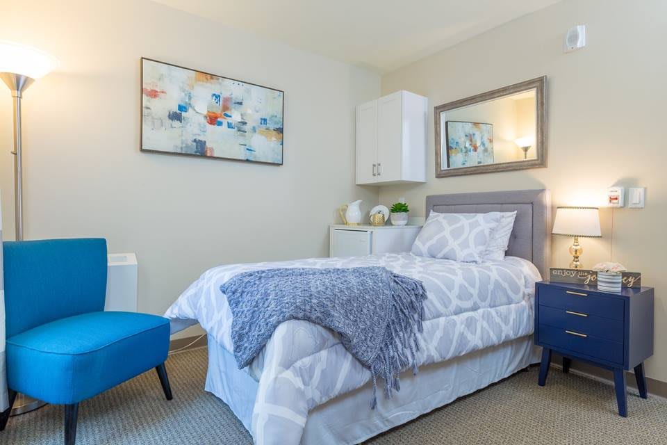 A modern bedroom with a single bed adorned with a geometric-patterned comforter and a blue throw, complemented by a blue chair and artistic wall decor.