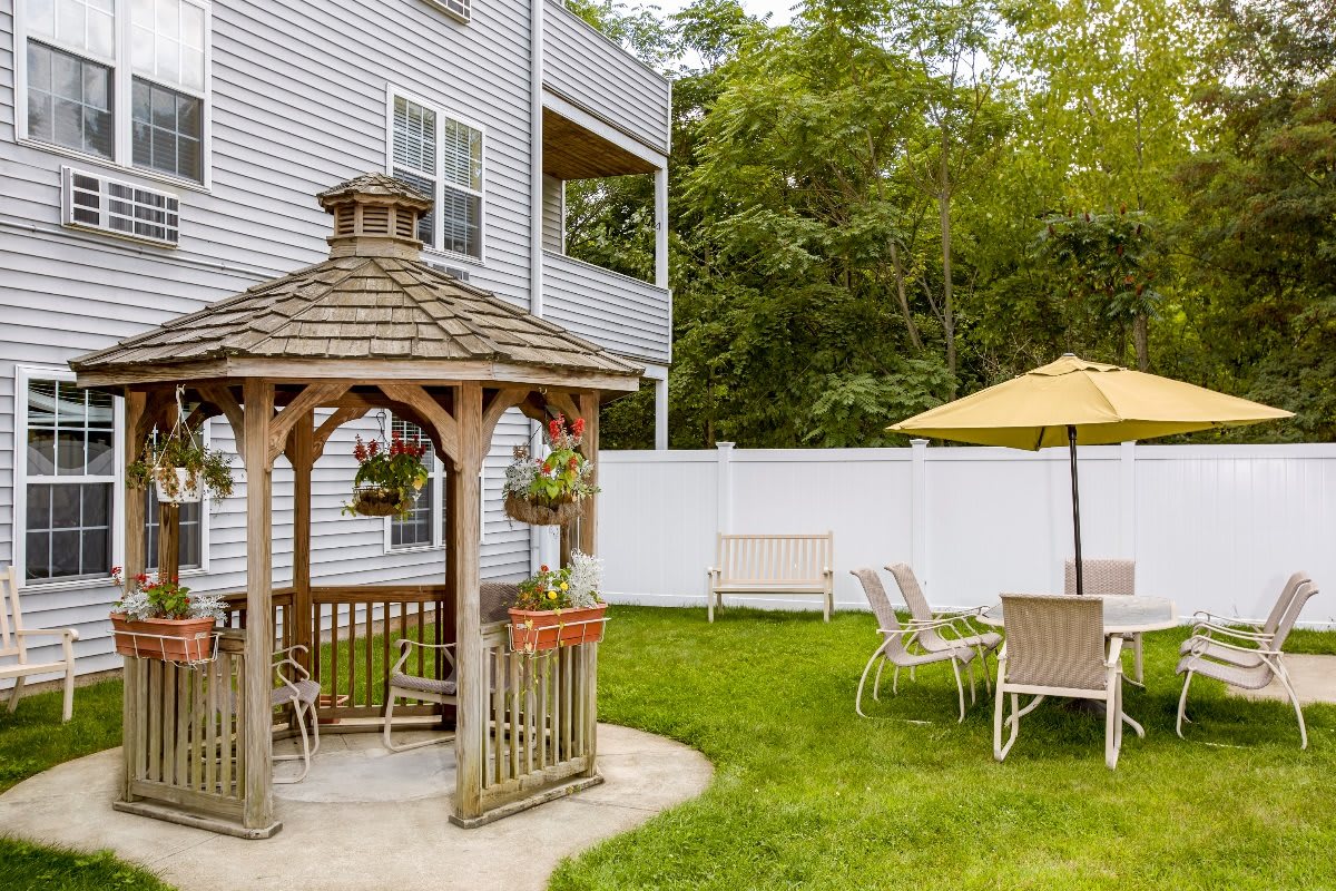 A cozy backyard featuring a wooden gazebo, outdoor seating with a yellow umbrella, surrounded by a white fence and lush greenery.