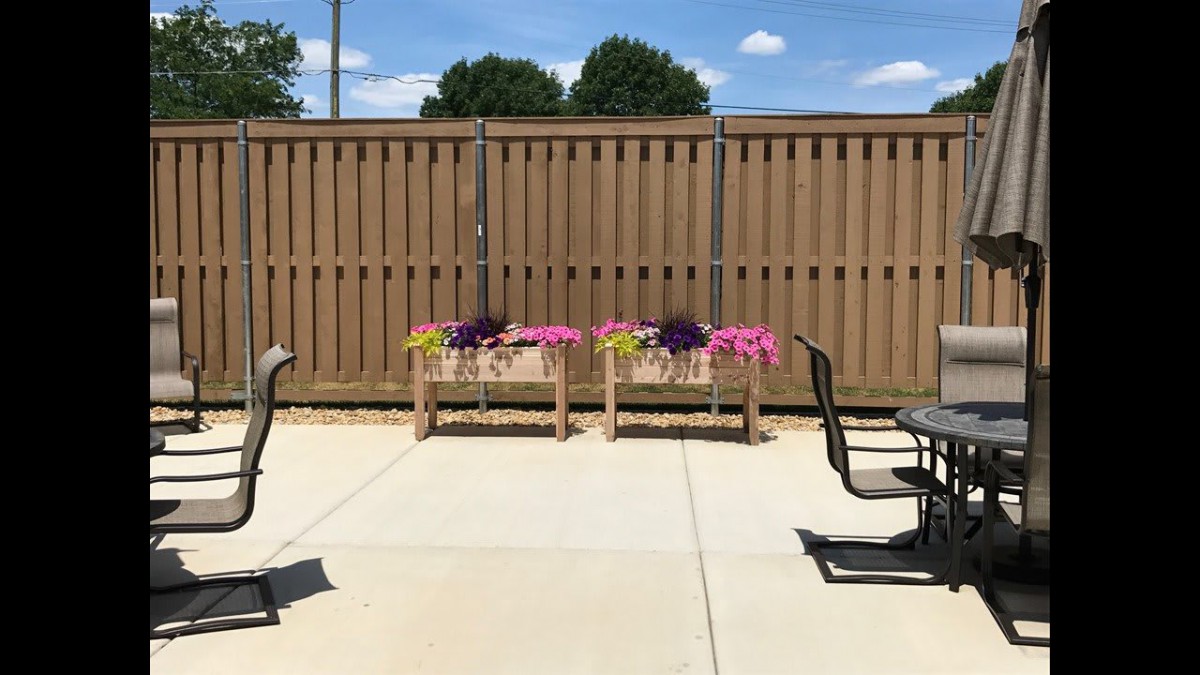 A sunny patio area with flower pots, chairs, and a table against a wooden fence.