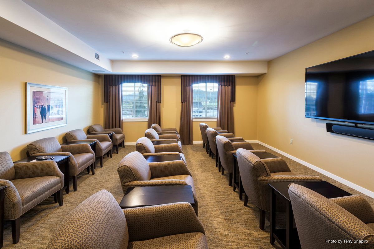 A small conference or meeting room with rows of comfortable chairs, natural light from two windows, and warm lighting.