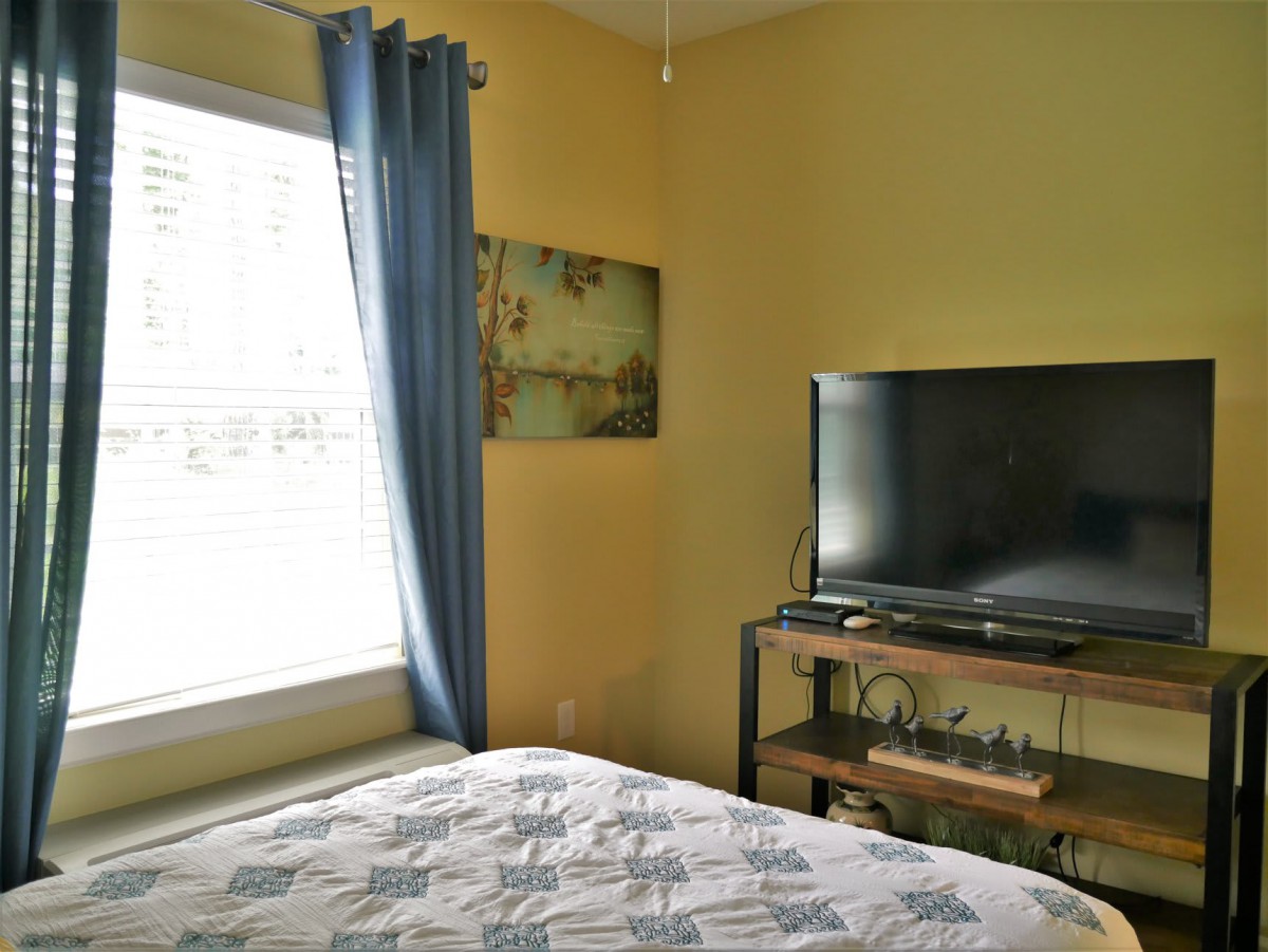A cozy bedroom with yellow walls, featuring a bed, a TV on a stand, and a window adorned with blue curtains.