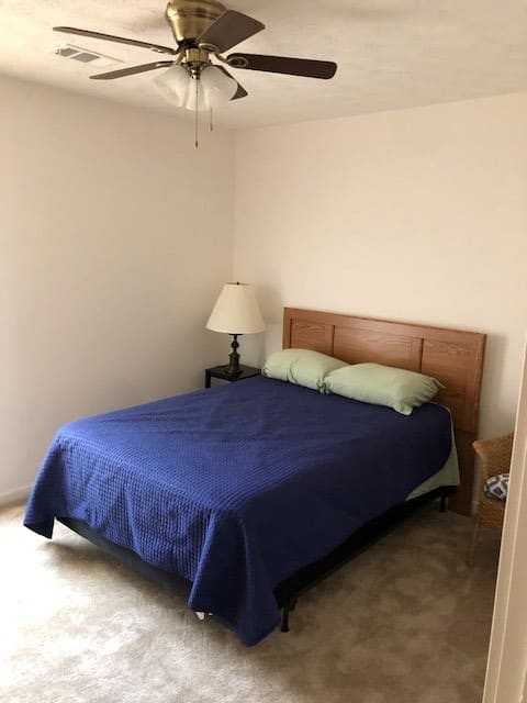 A simple bedroom with a bed covered in a blue comforter, two pillows, a lamp on a nightstand, and neutral walls.