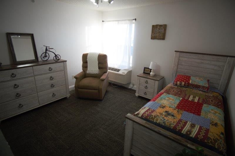 A cozy bedroom with a bed, armchair, and dresser arranged neatly under a bright window.