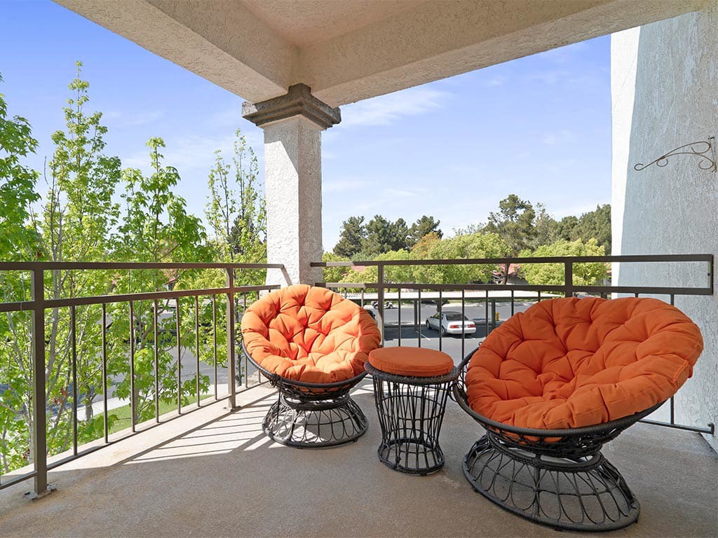 A sunny balcony with orange cushioned patio chairs and a table, overlooking a scenic view of trees.