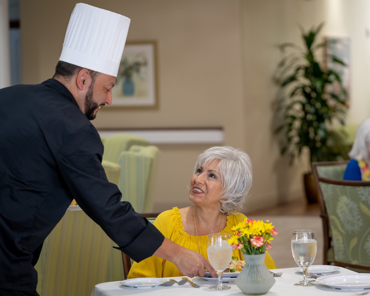 A chef serves a meal to a woman at a restaurant table.