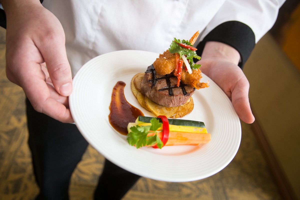 A chef presents a beautifully plated gourmet dish featuring beef, shrimp, and colorful vegetables.
