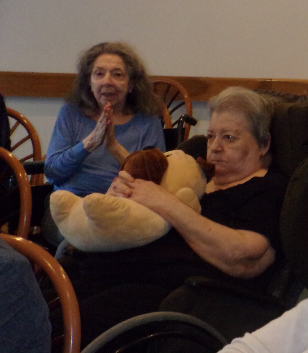 Two elderly women seated indoors, one holding a teddy bear.