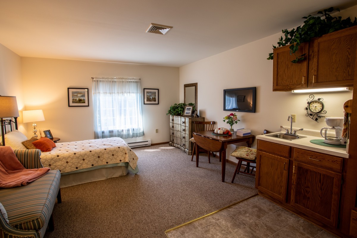 A cozy bedroom with a bed, desk, and dresser, featuring warm lighting and decorative elements.
