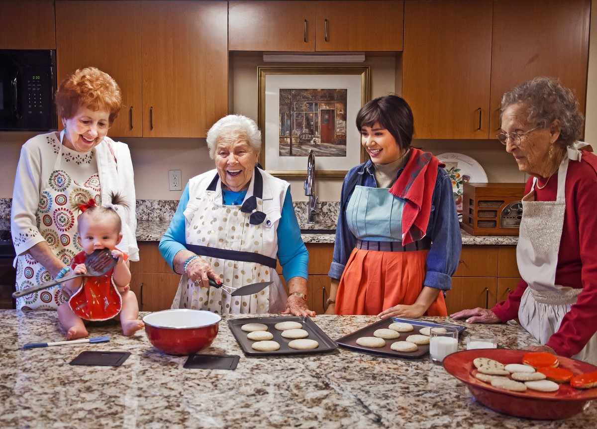 A group of people baking cookies together in a kitchen, all wearing aprons and smiling.