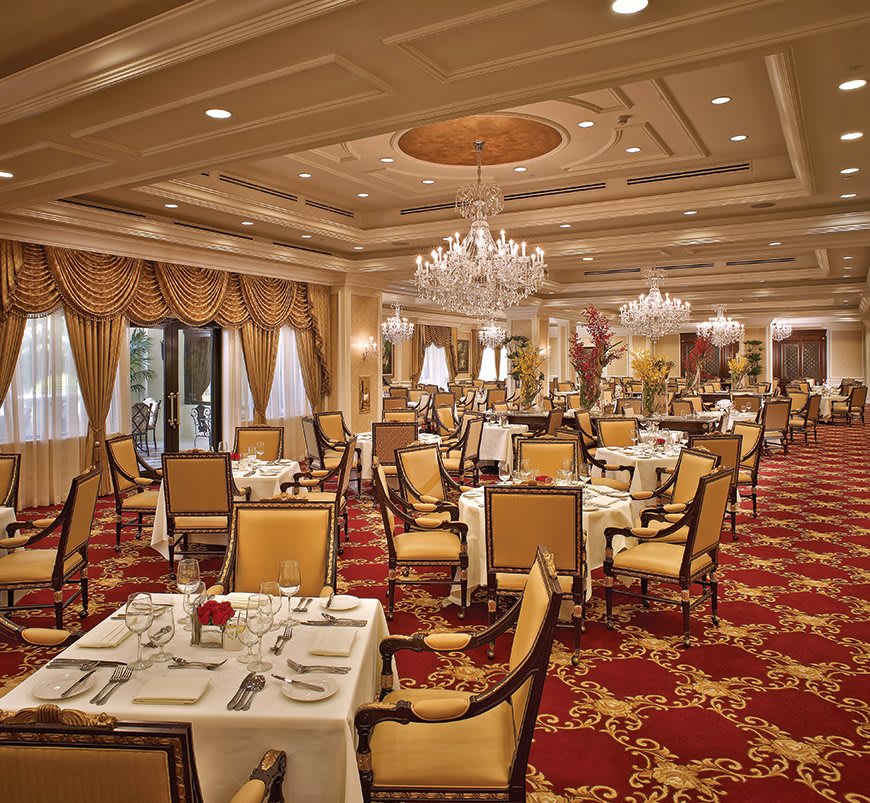 A luxurious dining room adorned with chandeliers and elegant decor, featuring neatly arranged tables and chairs on a richly carpeted floor.