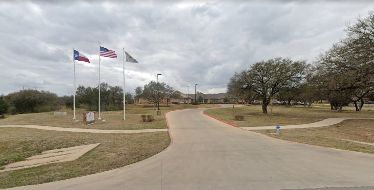 A paved road leading to a building with flags and trees under a cloudy sky.