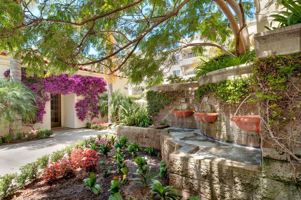 A tranquil garden featuring vibrant flowers, a stone wall, and lush greenery under the dappled sunlight.