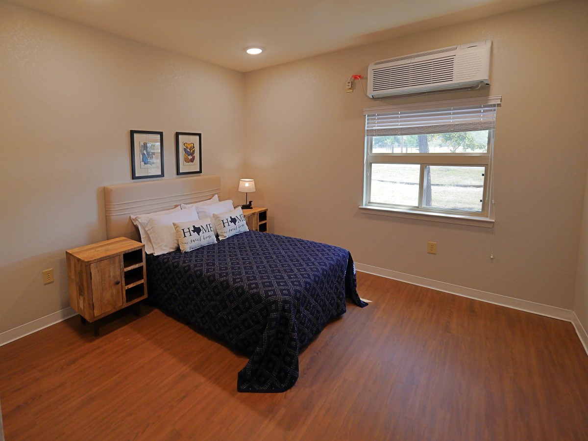 A minimalist bedroom with a neatly made bed, wooden flooring, nightstands, and wall art. The room is well-lit with natural light coming through the window.