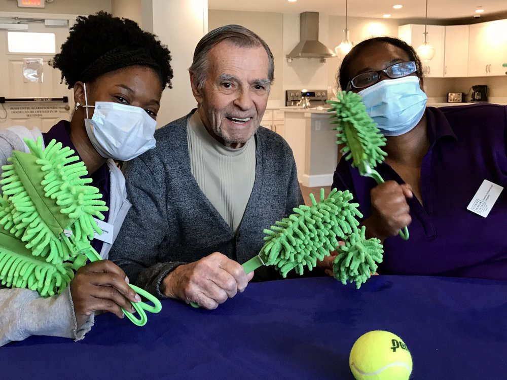 A group of two healthcare workers and a senior individual sitting together, all smiling and wearing masks indoors.