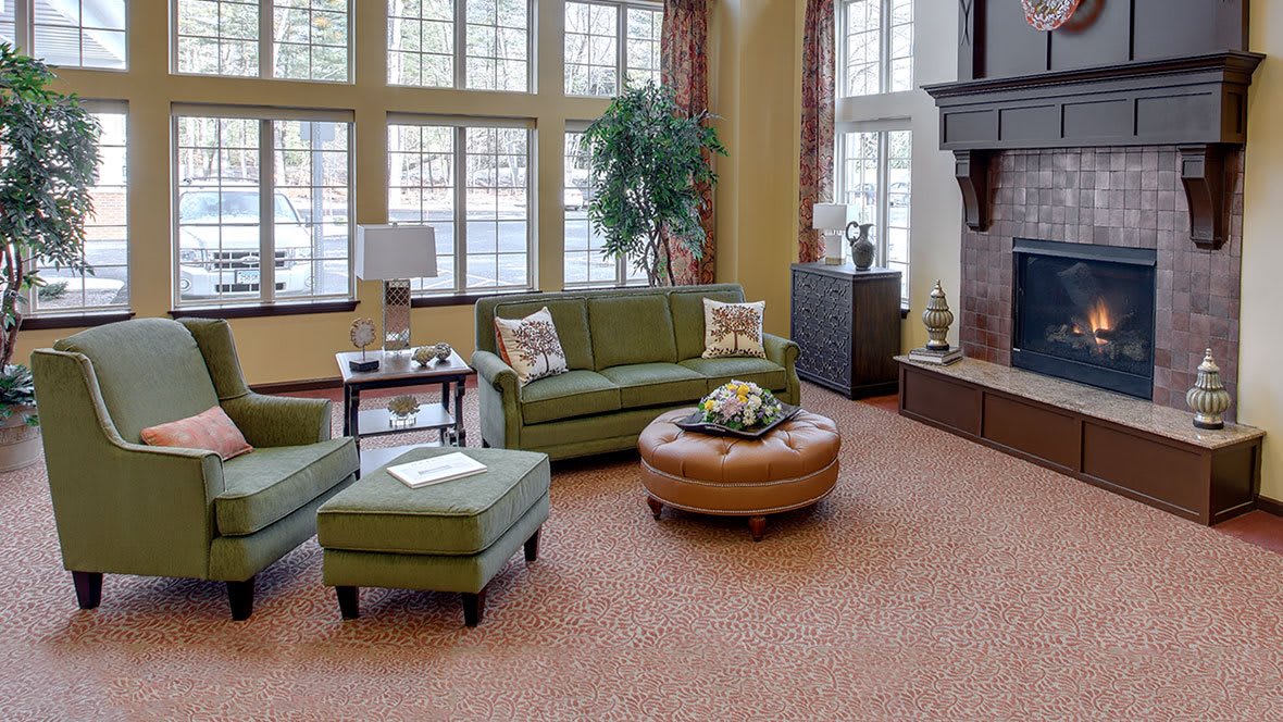 A cozy living room with green sofas, a brown ottoman, and large windows.