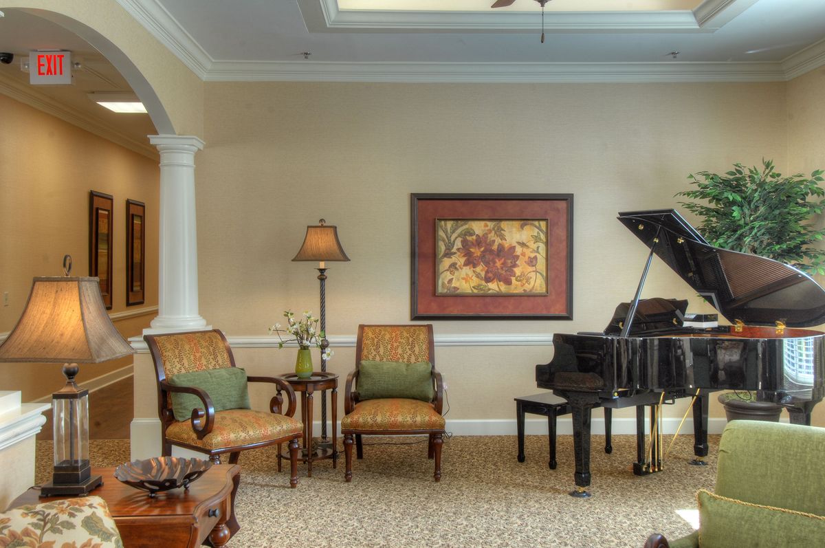 A cozy living room featuring a grand piano, two chairs, a lamp, and framed artwork on the wall.