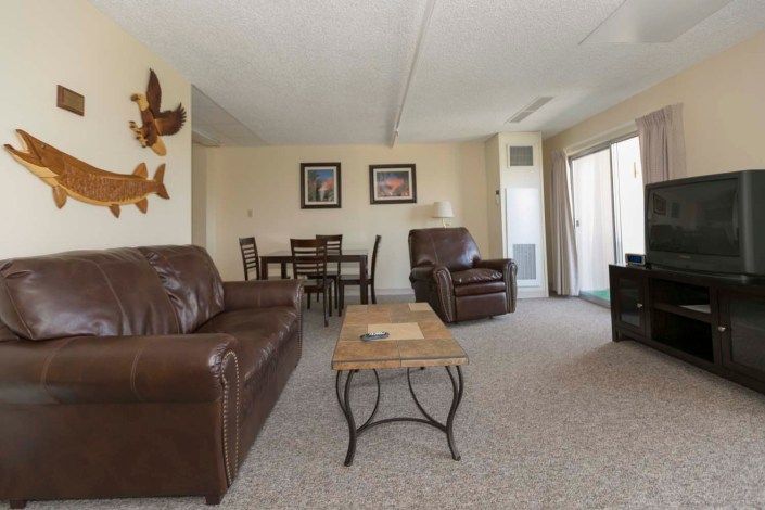 A spacious living room with comfortable leather seating, a wooden coffee table, and a dining area in the background.