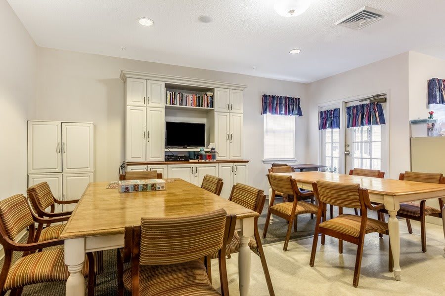 A well-lit dining room featuring wooden tables and chairs, a television mounted in a cabinet, and two windows with blue curtains.