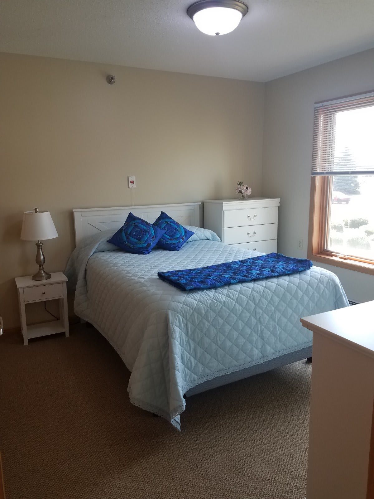 A cozy bedroom with a neatly made bed, side table, lamp, and dresser near a window.