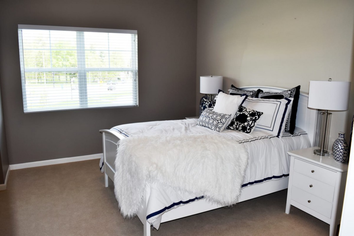 A cozy bedroom with neutral tones, featuring a bed with decorative pillows and a lamp by the window.