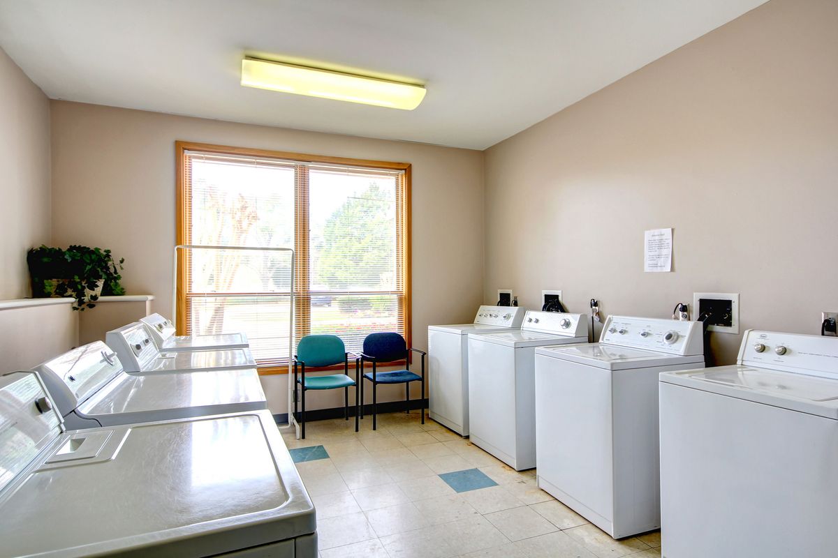 A clean and bright laundry room with several washing machines and dryers along with chairs by a large window.