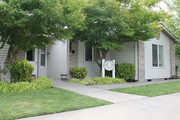 A residential building with a landscaped front yard, featuring trees and bushes along a concrete sidewalk.
