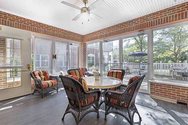 A cozy sunroom with wicker furniture, surrounded by brick walls and large windows, featuring a ceiling fan for comfort.