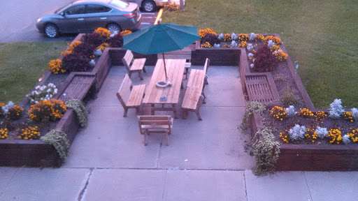 A cozy outdoor seating area with a wooden table, chairs, and a green umbrella surrounded by flowers.