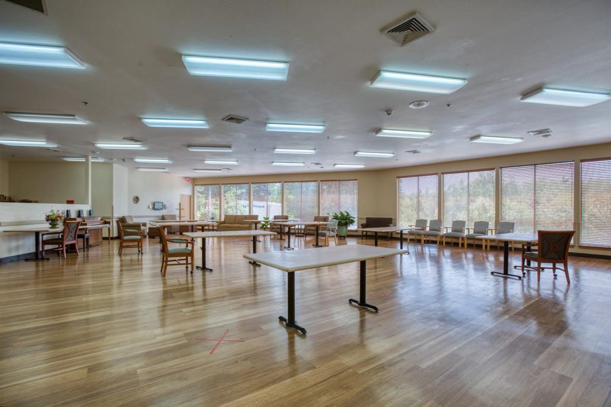 An empty community center room with tables and chairs arranged on a wooden floor, illuminated by fluorescent lights.
