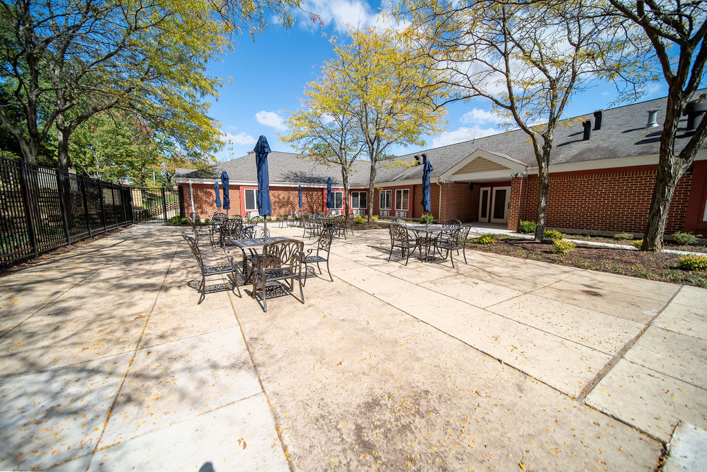 A sunny outdoor patio with tables and chairs, surrounded by brick buildings and trees.