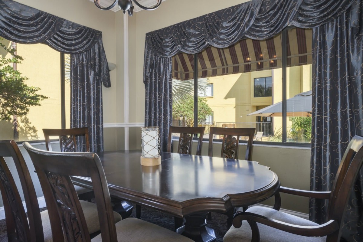 A dining room featuring a large wooden table and upholstered chairs, with windows adorned by dark curtains.