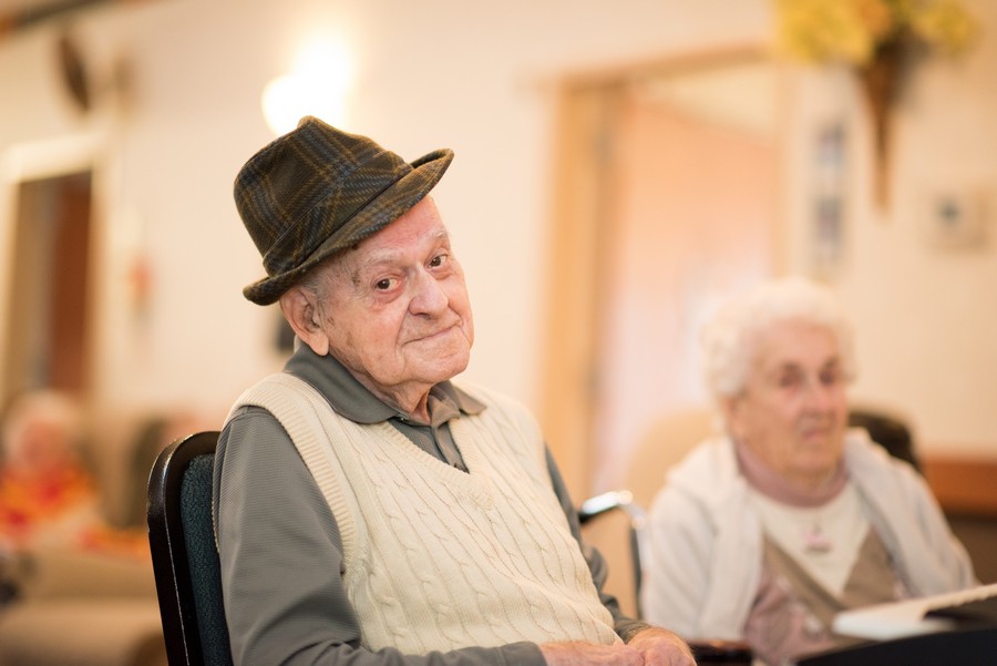 An elderly man wearing a hat sits indoors, with a woman in the background.
