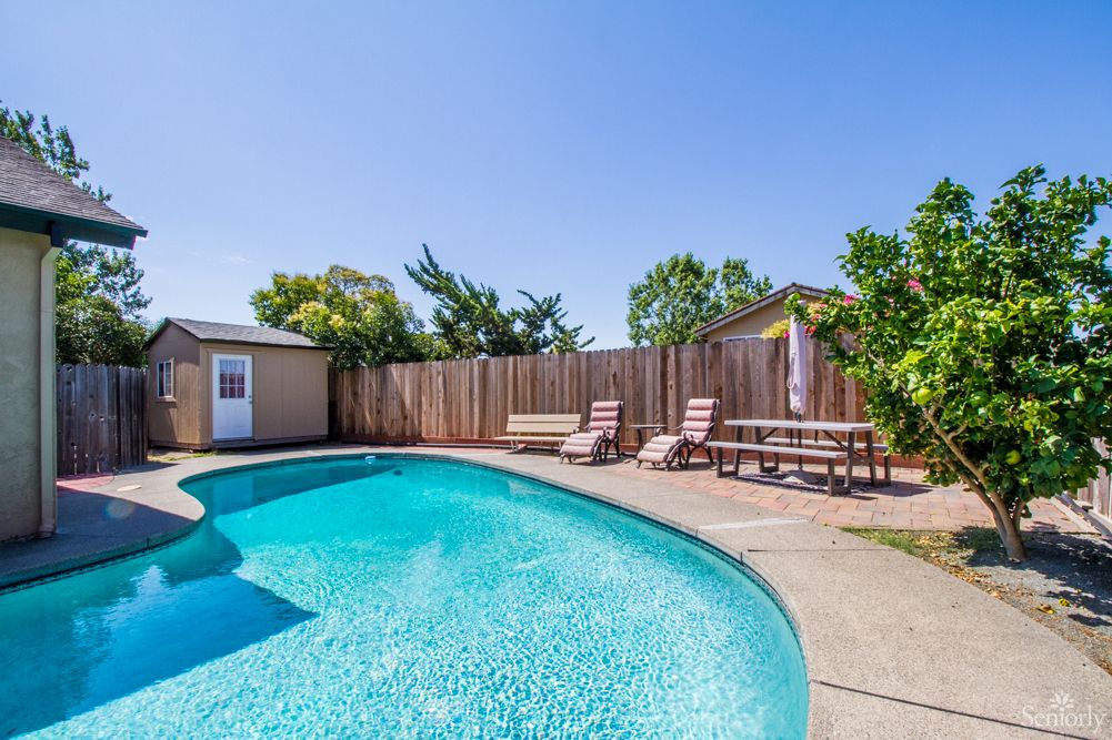 A sunny backyard featuring a swimming pool with lounge chairs and a wooden fence.