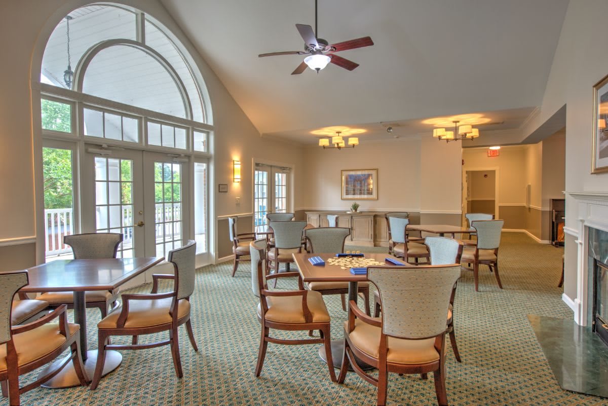A well-lit dining area with tables and chairs positioned under a ceiling fan and large windows.