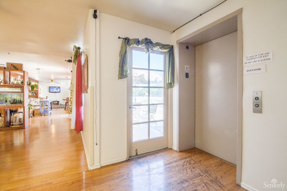 A residential hallway featuring an elevator and a door with curtains, all on a wooden floor.